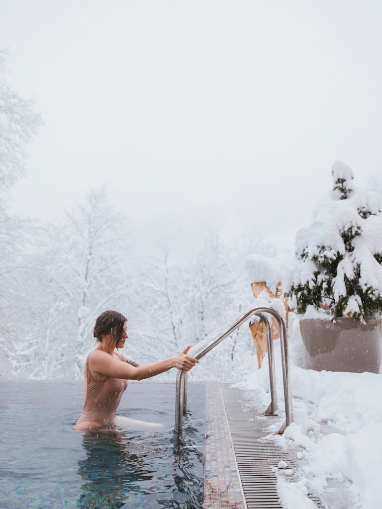 A woman in a swimsuit enters a heated pool surrounded by snow-covered landscape in winter.