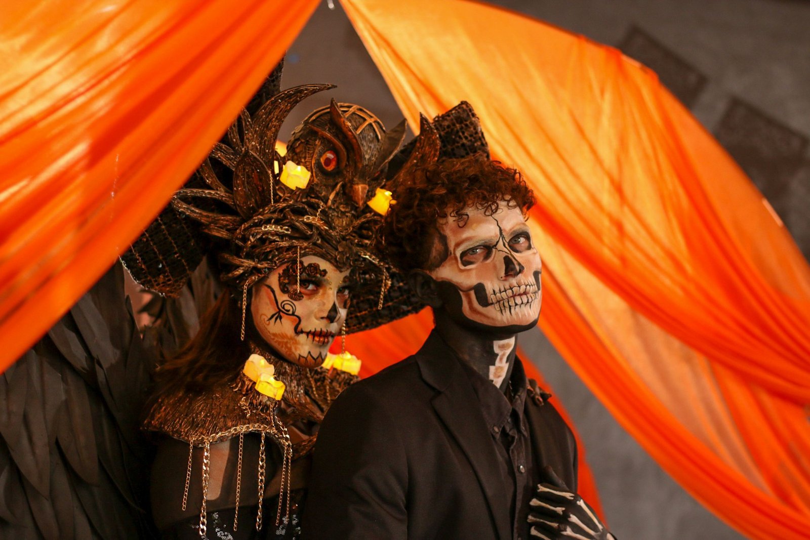Striking Dia de los Muertos couple in elaborate skeleton costumes with vibrant orange backdrop.